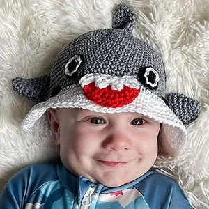A baby is lying on their back on a white fluffy carpet and looking just passed the camera. They are wearing the grey shark with white brim. It has a fin at the crown and 2 at the sides, a red mouth with white teeth and two black eyes.