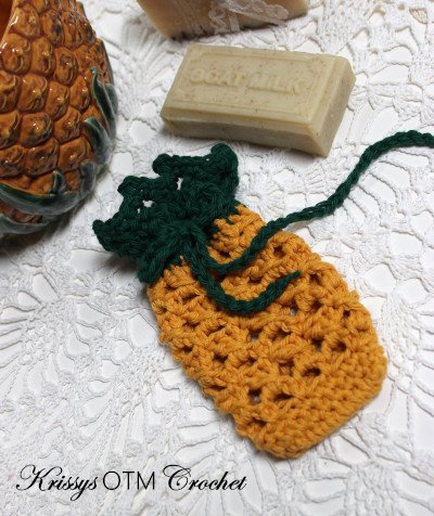 A pineapple shaped and styled crochet soap saver. The body of the bag is yellow and has a pattern similar to a pineapple. The top drawstring is green and shaped like pineapple leaves. The background is white and there is a cream bar of soap above.