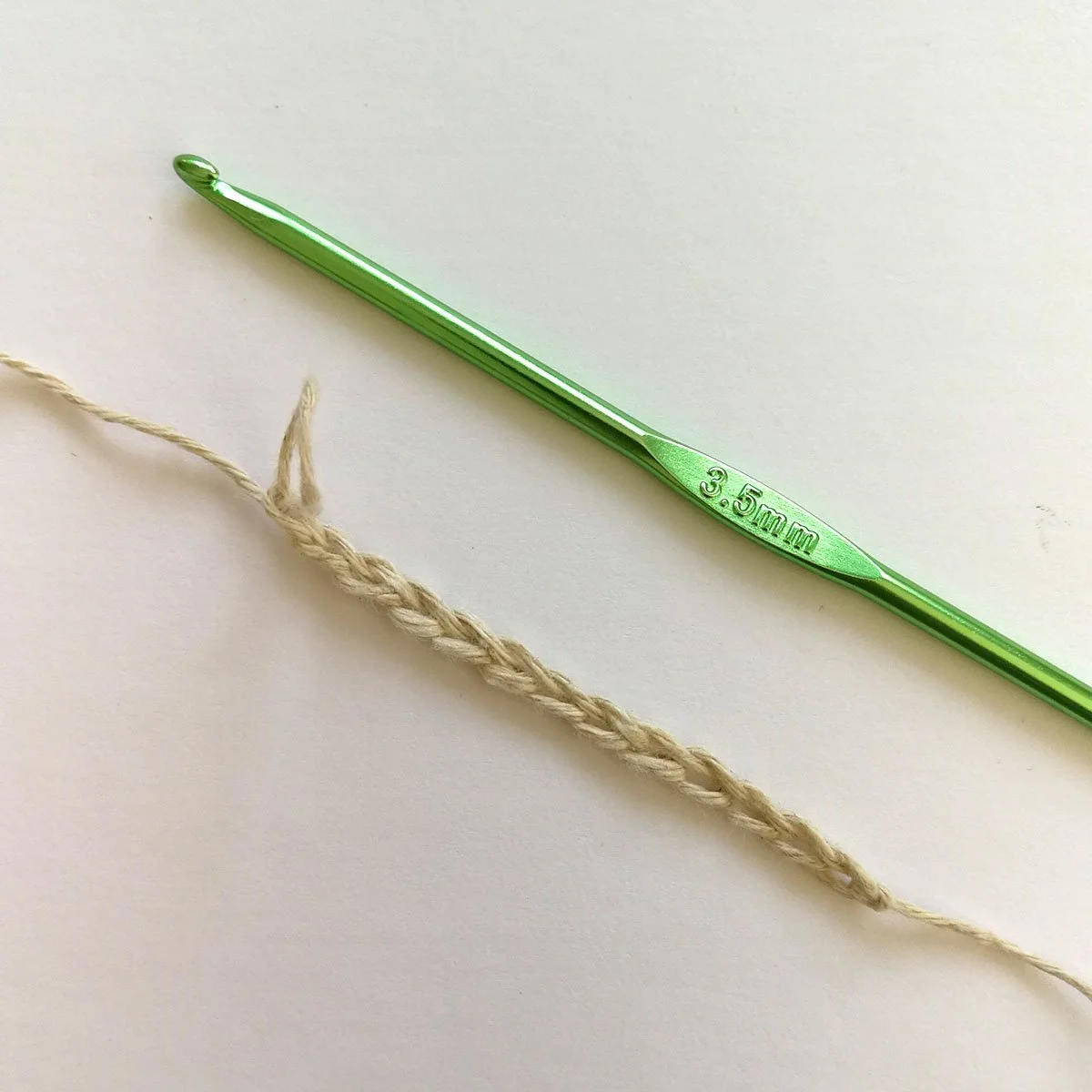 A small foundation chain of 12 chains lying diagonally across from top left to bottom right. A bright green crochet hook is lying parallel with the hook pointing towards the top. 3.5mm is written in the middle of the hook on a flat section.