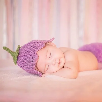 A baby is laying on their front with their head turned to the left facing the camera. They have their eyes shut like they're sleeping. They are wearing a rose coloured tulip hat with a green stem on the crown. The background is soft pink.