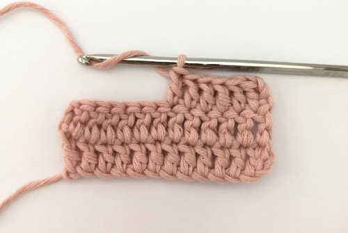 A small sample of crochet work worked in a pale pink yarn. There are two full rows of 12 double crochet stitches and half a third row. The working yarn is wrapped over the silver crochet hook ready for the next stitch.