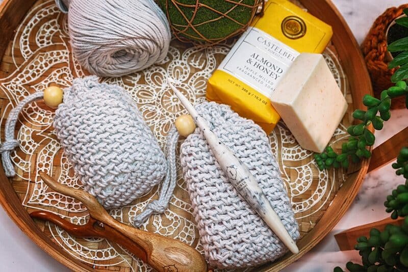 Two light grey soap bags inside a round wooden serving tray with a white doily styled crochet piece in the bottom. The soap bags has a simple even stitch with a drawstring close and a round wooden toggle.