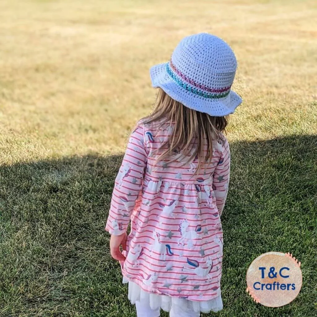 A young girl is standing on green grass. She is wearing a pink dress and the criss cross bucket hat which is white with two different coloured lines around just above the brim. She has shoulder length light mousey coloured hair.
