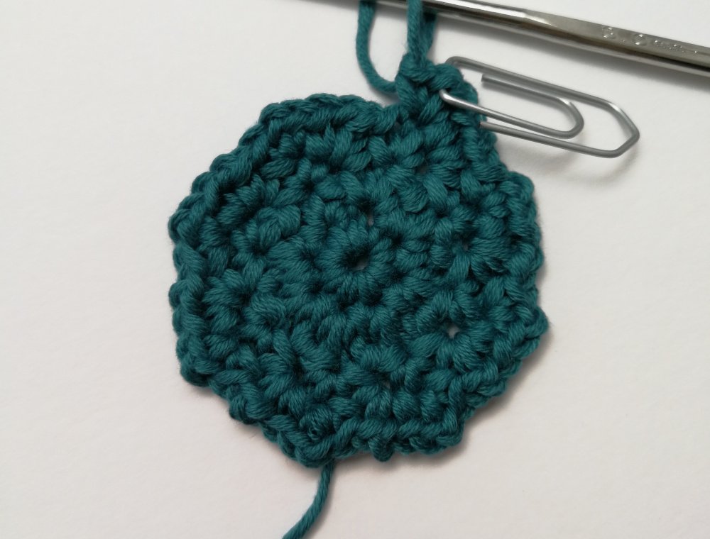 A close up photo of the first three rounds of the crochet face scrubbie. It is made in teal yarn, has an uneven edge all the way around and a silver paper clip marking the last chain 1. There is a silver crochet hook attached to the loop.