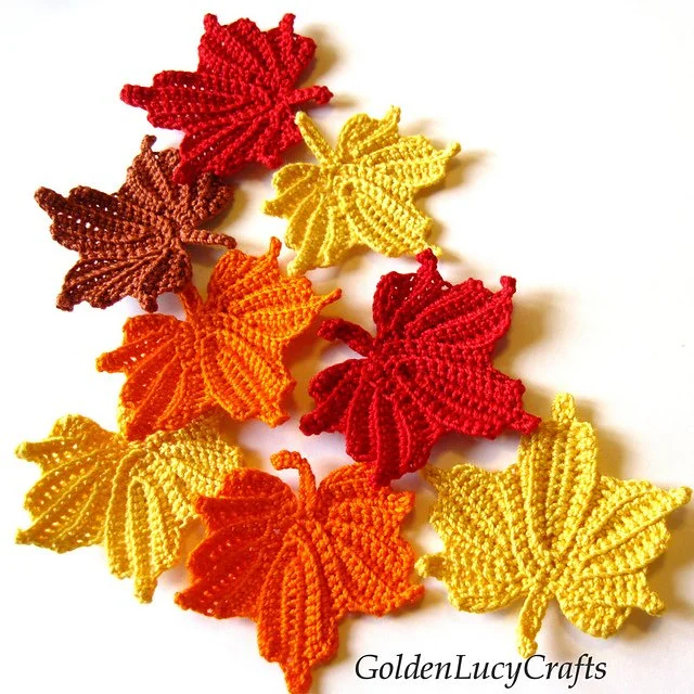 A white surface with 8 small, crocheted maple leaves in autumnal colours red, yellow, orange and brown.