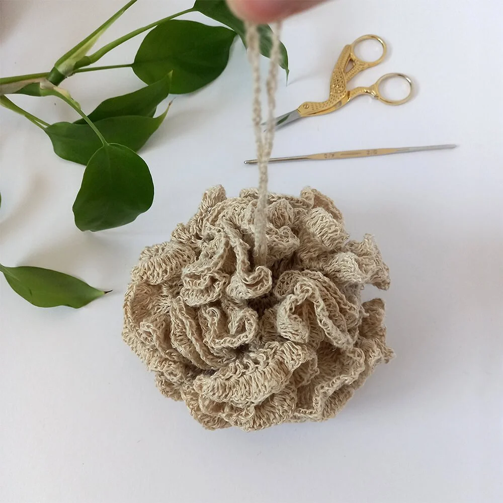 A birds eye view of a beige crocheted loofah that looks just like a store bought, mesh, shower pouf. The hanging loop is being help up so it is straight up. In the background there are green leaves and a metal pair of scissors and crochet hook.