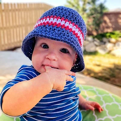 A baby is sat outside, facing the camera, nibbling at their thumb. They are wearing a blue and white striped top and a dark blue bucket hat with a red and white stripe around the body.