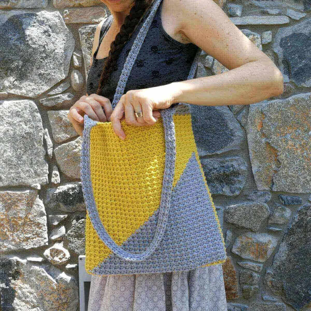 A slim white person is facing left stood in front of a grey stone wall. They have one strap of a yellow and grey crocheted tote bag over their shoulder and they are looking inside the bag. Their head is not visible.