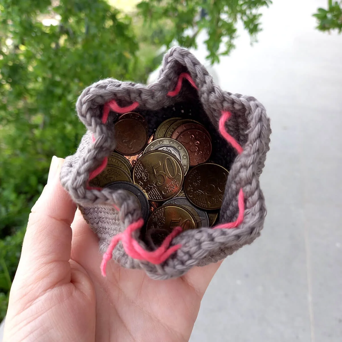 A birds eye view of the open, warm grey coin purse sat on a white left hand. The opening is shaped into a 6 pointed star. About two rows down there is a bright pink drawstring weaved all around. Inside the purse is a mix of coins.