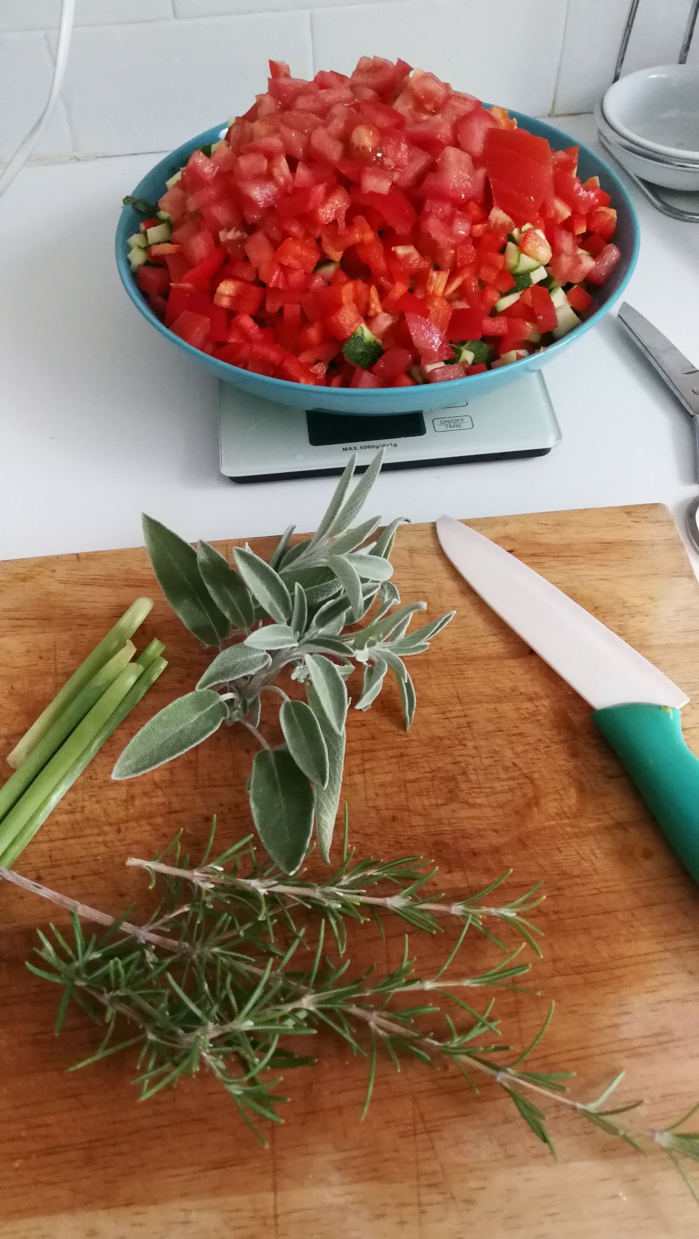 A photo of a white kitchen work surface. A wooden chopping board has sage, rosemary sprigs and spring onions on it with a green and white knife. In the background is a blue bowl on top of a digital scales full of chopped tomatoes, peppers, courgettes