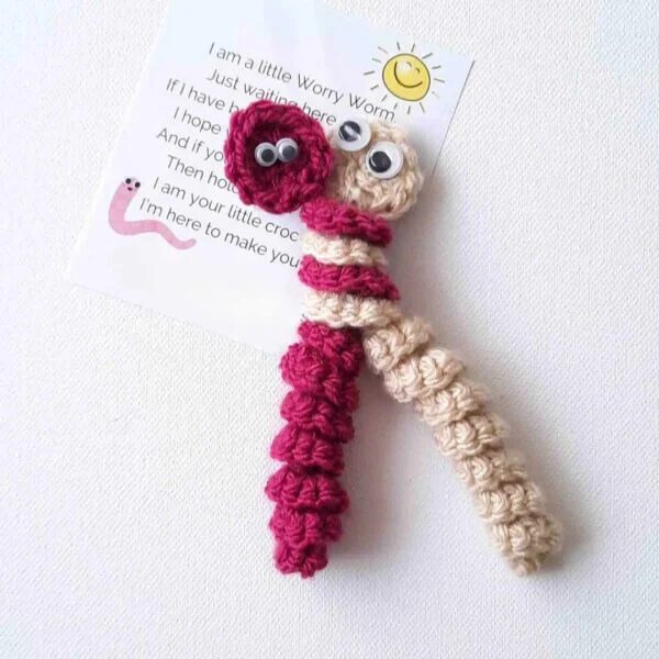 A birds eye view of two worry worms with their top sections entwined together. The left one is magenta and the right is cream. They both have a small circle as a head, googly eyes and a spiral body. The background is white.
