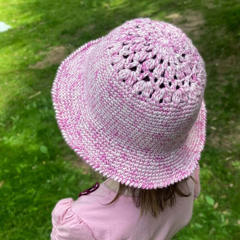 A young person with mousy brown shoulder length hair is facing away from the camera towards the left top corner. They are wearing a pink cotton top and a pink and white crocheted hat. The hat has a floral pattern around the crown and a sturdy body.