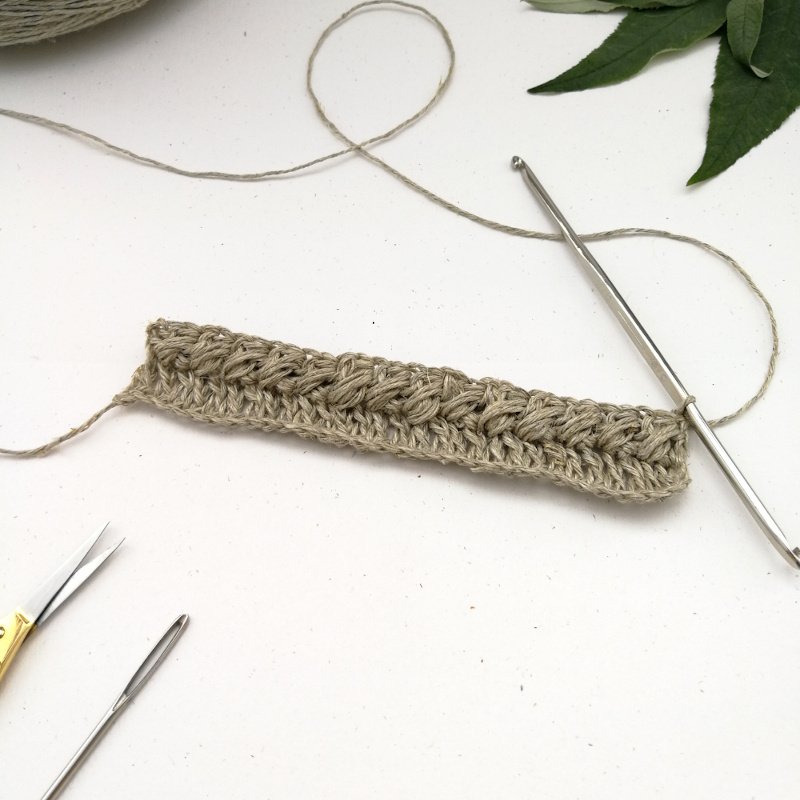 A small sample of crochet work is sat on a white background. There are two rows of crochet, one simple stitch and one puff stitch. The silver crochet hook is still attached to the last loop.