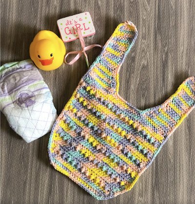 A multicoloured bib is sat on a dark wooden block next to a nappy, a yellow duck and a "It's a Girl" tag. The bib has bobble stitches all across the front and is made up of yellow, blue, pink colours.