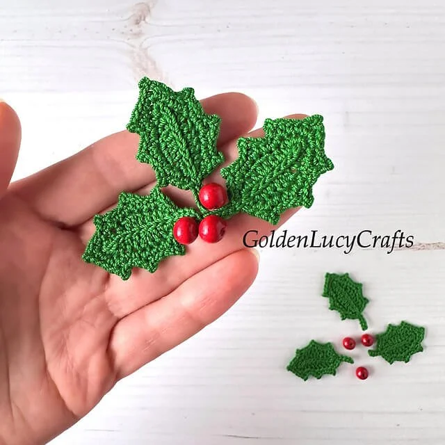 A birds eye view of a left white hand holding three crocheted, green holly leaves in their hand with shiny red berries in the centre.  The background is off white.