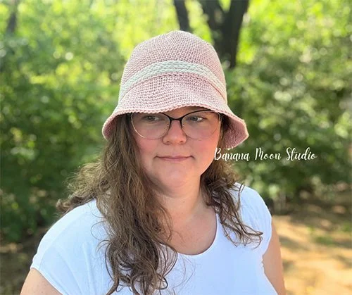 A white person with long wavy brown hair is facing the camera. She is wearing the Summit hat which is light peach in colour and has a white band around the body just above the brim. The brim is medium in length and sits close to the face.