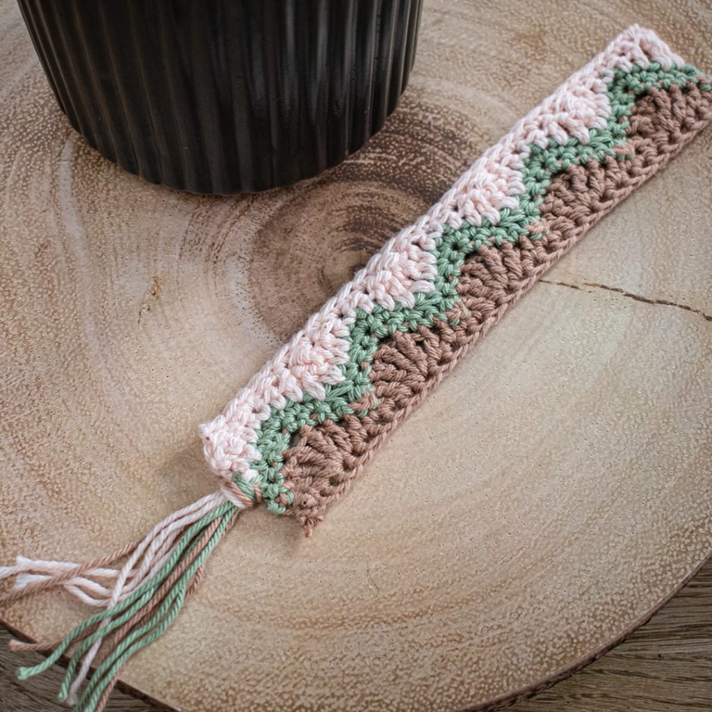 A zig zag patterned bookmark is sat on a wooden block. It is split in two with the left side being cream, the right side light brown with a green zig zag horizontally through the middle. All colours are brought together into a tassel at the bottom.