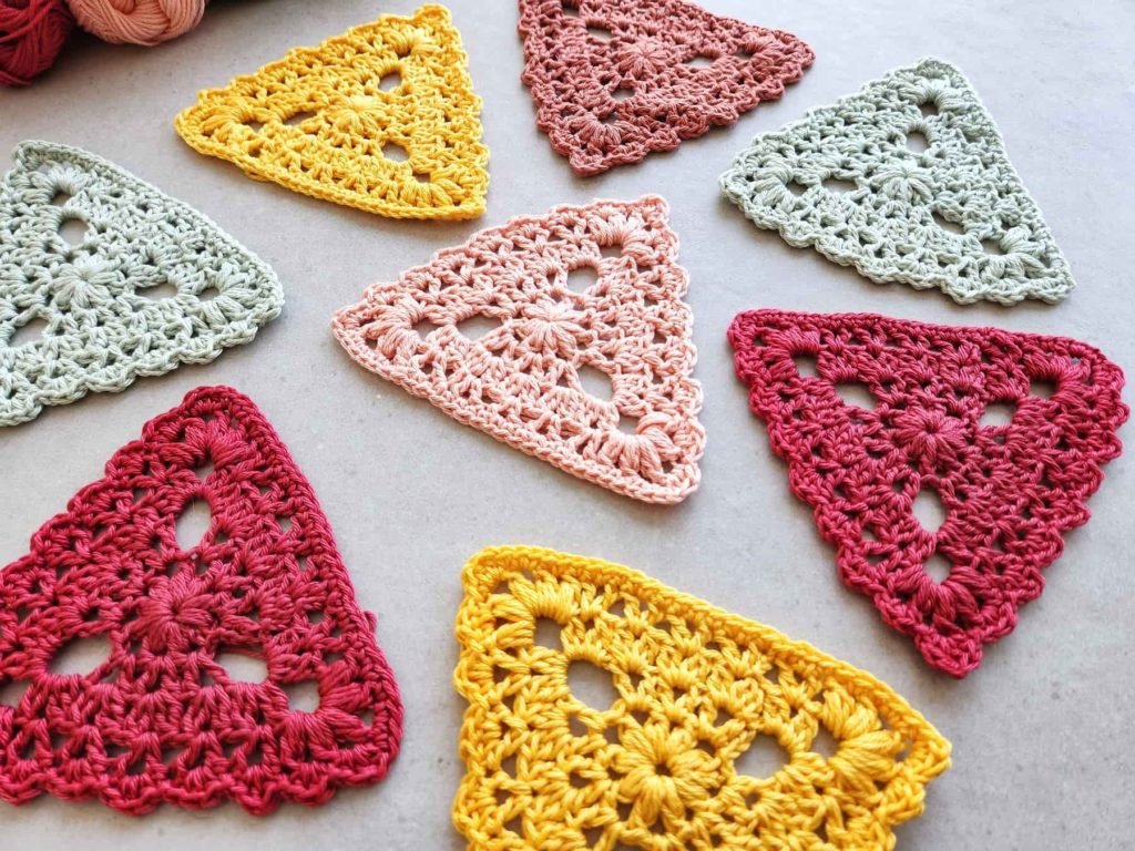 Eight small crochet triangles are arranged randomly on a light grey table. The colours are yellow, magenta, light green, light pink and dark pink.