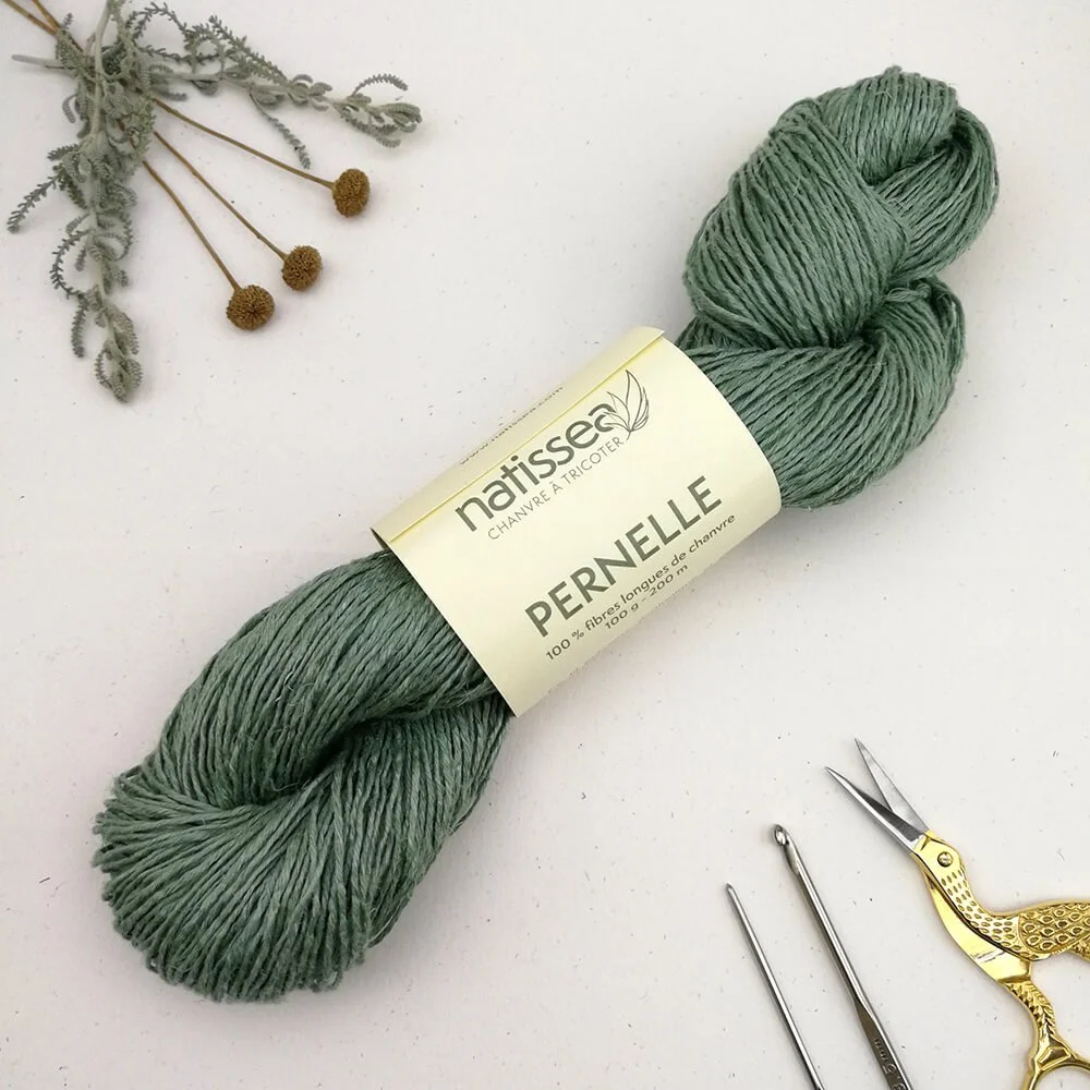 A birds eye view of a pastel, green skein of hemp yarn laying diagonally across a white surface. It has a cream coloured paper band around its middle with green text that reads "Natissea" and "Pernelle". There are lavender cotton flowers at the top.