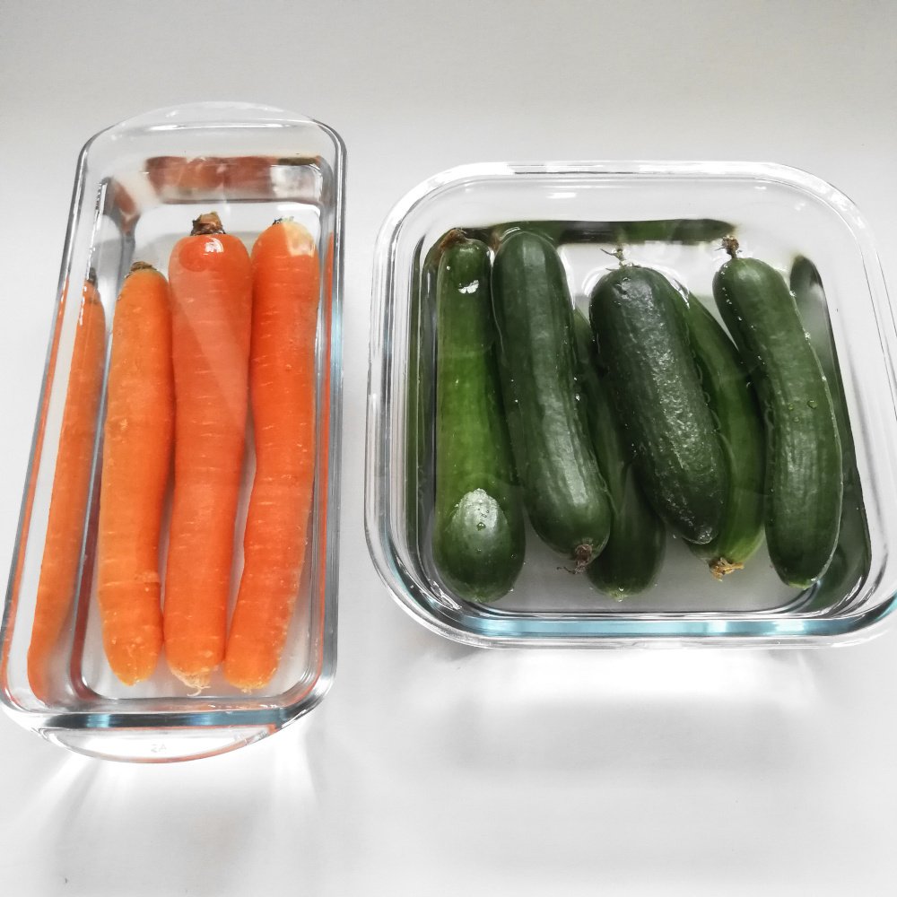 A rectangular glass dish and a square glass dish are sat on a white counter top side by side, left to right respectively. There are 3 carrots in the rectangular dish and a 6 cucumbers in the square dish.