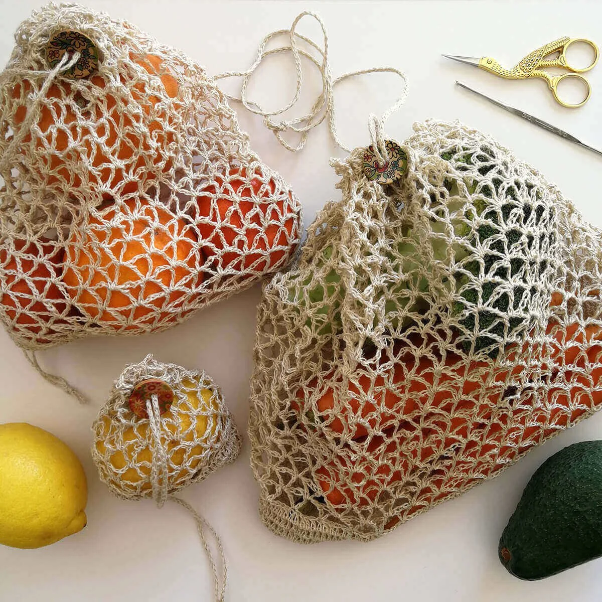 A birds eye view of three different sized crocheted produce bags. They have been made in a beige coloured yarn and the design is a net with V shapes. They have a drawstring top with a wooden button to close. The background is off white.