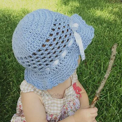 A baby sat in green grass holding a stick and wearing a white and cream checkered dress. They are wearing a blue crocheted summer sun hat with a solid crown, open weave body and slightly ruffled brim. There is a white ribbon round the end of the body