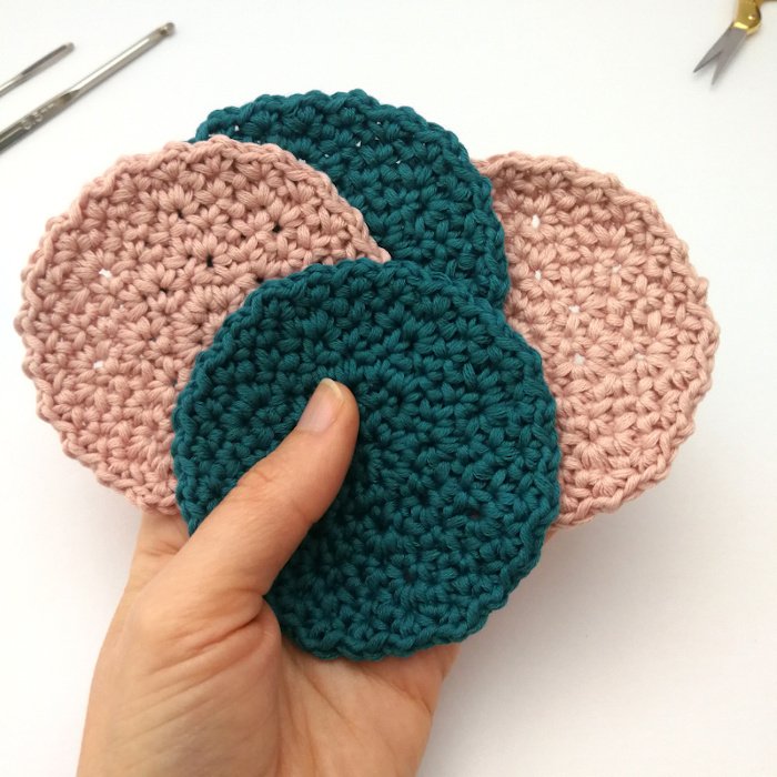 A white left hand is holding four round, crocheted facial scrubbies like a hand of cards. Two are teal in colour and the other two are pastel pink. They are flat with a level, textured surface and an uneven edge. The background is off white.