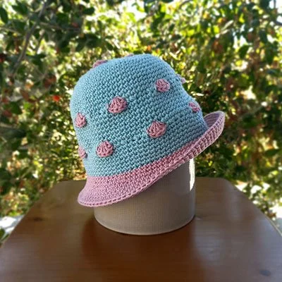 A small bucket hat placed on top of an upside down, grey plant pot, facing to the right. The body is egg shell, the brim is rose and there are rose shells crocheted around  the body. The brim is turned up on at the front, facing right.
