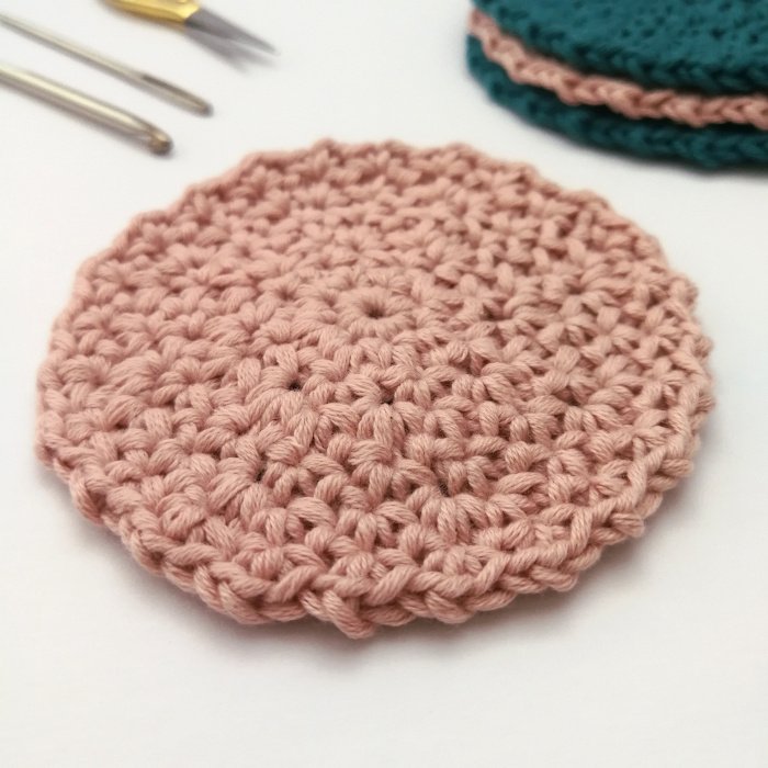 A close up photo of one of the pastel pink crochet face scrubbies. It is round and has an uneven edge all the way around with a smooth surface and a small hole in the centre. The background is off white,