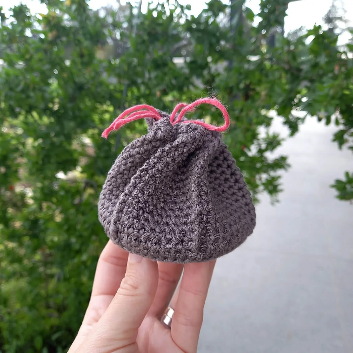 A warm, grey coin purse perched on top of white fingers. It is a small hexagonal pouch with a round base, twisted closed at the top with a drawstring that pulls into a twist.