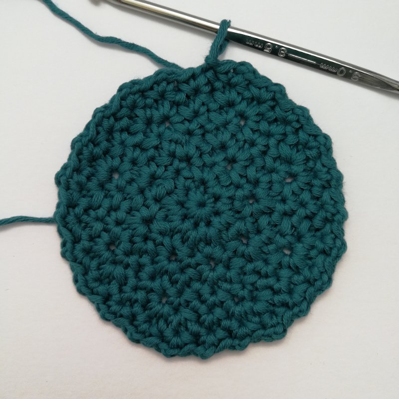 A full sized face scrubbie made in teal yarn. It has six rounds of stitches and an uneven edge. The silver crochet hook is still in the last loop