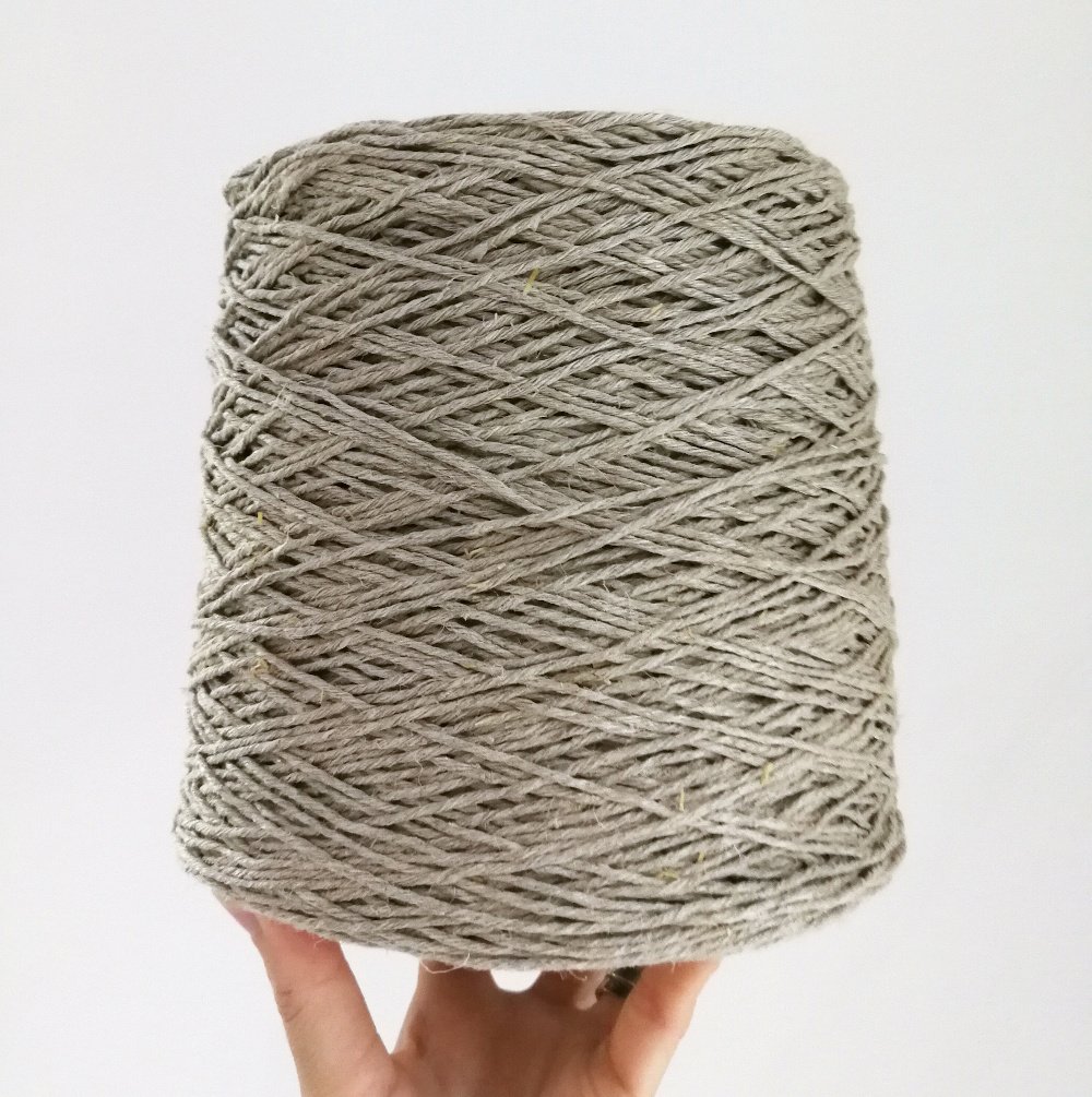 A left hand is holding up a one kilo cone of hemp yarn in DK weight. The cone is balancing on the hands fingertips. The yarn is light grey in colour and the background is off white.