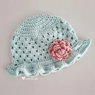 A pastel duck egg crocheted hat with a pink flower is laying flat on a grey surface. The crown is solid with an open weave body and a ruffled brim.