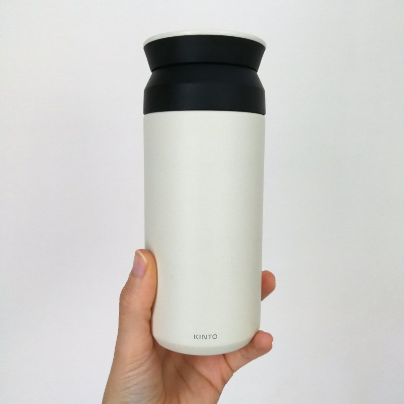A white left hand is holding up a small off white flask with a black lid. The background is light grey.