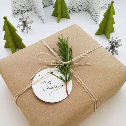 A square gift wrapped in brown paper and tied with natural coloured twine with a bow in the centre is sat on a white surface at an angle. There is rosemary under the bow and a round white card with the words "Merry Christmas" written in black.