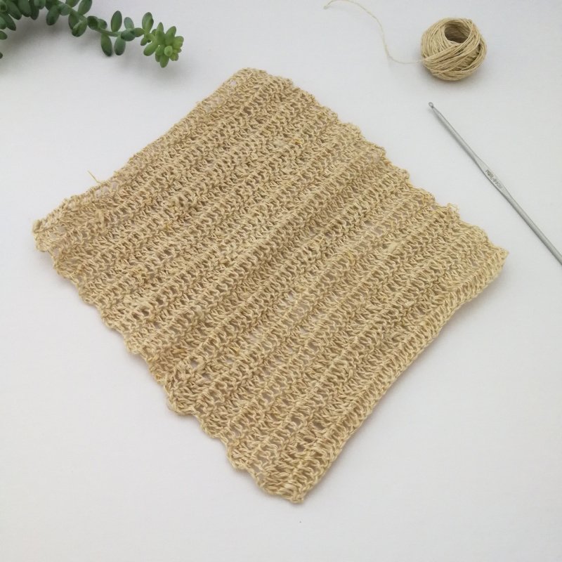A neutral coloured hemp dishcloth is laid out in a diamond shape on a white table. It has a simple stitch which is repeated on each row. There is a grey crochet hook on the right next to a small ball of yarn. There is a succulent in the top left.