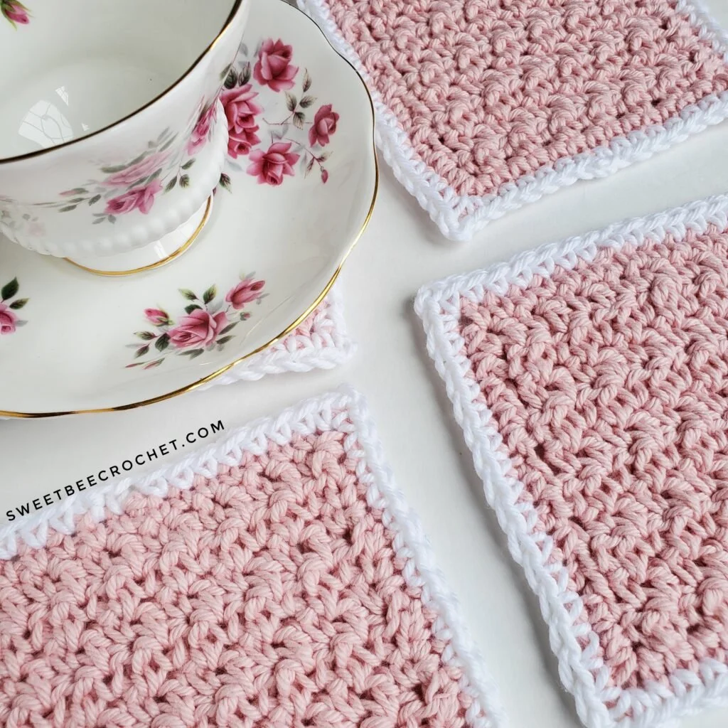 A birds eye view of 4 square coasters made in light pink yarn with a white border. A white teacup and saucer with pink flowers is sat on the top left coaster. They are all half in the photo and half out and angles.