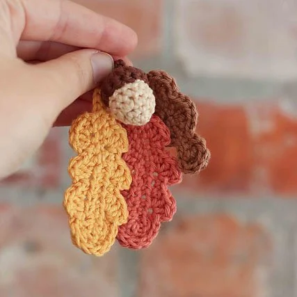 A left hand is holding up three crocheted oak leaves in yellow, red and brown with a small cream and brown acorn in the centre. The background is a blurred brick wall which adds to the autumnal feel.