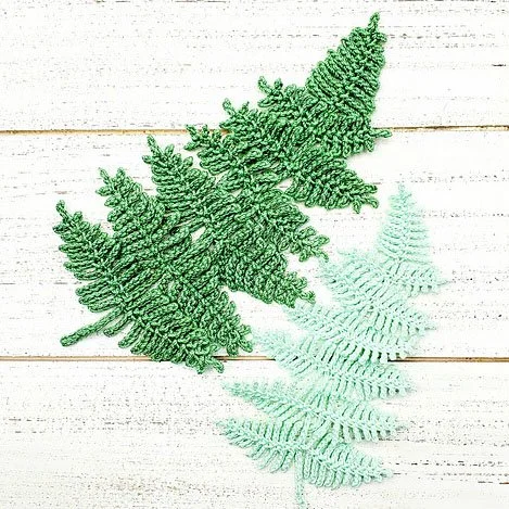 A birds eye view of two fern leaves made with crochet. They are laying on a rustic white, wooden background. One is true green and the other small one is light, pastel grey green