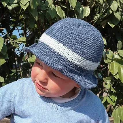 A toddler is sat in front of a green bush wearing a blue v-neck top and the derby summer hat which is also blue with a white stripe across the body just above the brim. The brim is short and wavy but sturdy.