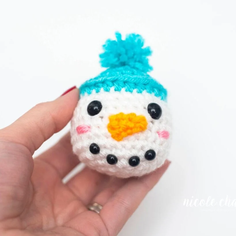 A white left hand with red nail varnish is holding a round snowman head up to the camera and against the white background. The head has a light blue hat with a bobble onto, two black eyes, an orange carrot nose, rosey cheeks and a black dotted smile.