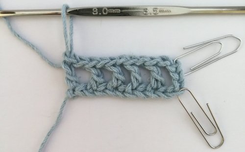 A photo showing 5 double crochets done in baby blue yarn with the chain 3 on the right, so 6 in total with gaps in between. Two silver paper clips as stitch markers and the silver crochet hook still attached to the working loop.