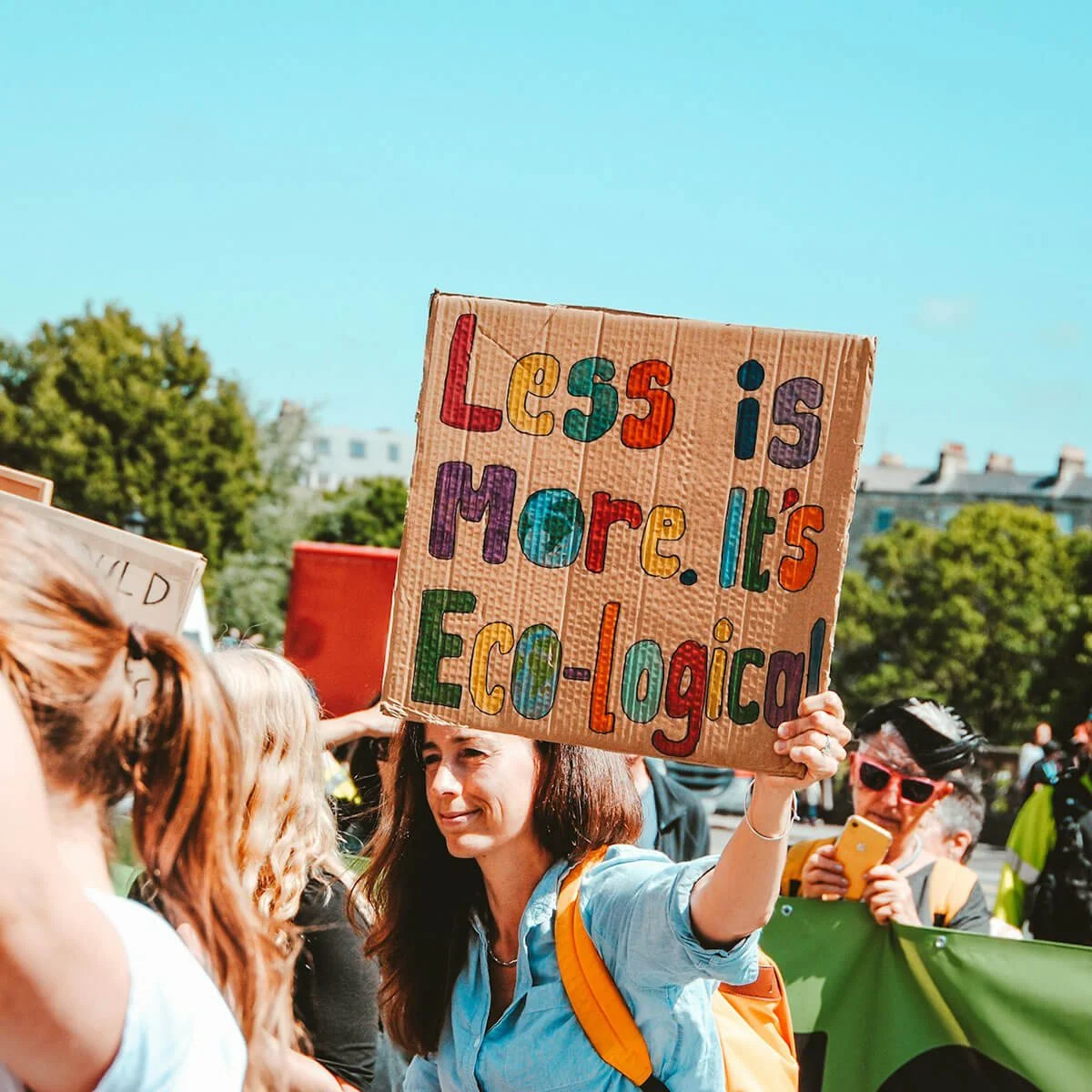 People at a demonstration holding cardboard cut outs with written text. The main focus is a smiley brunette with medium length hair and a light blue shirt. She is holding a sign that says Less is More. It's Eco-Logical. Behind are trees and sky.