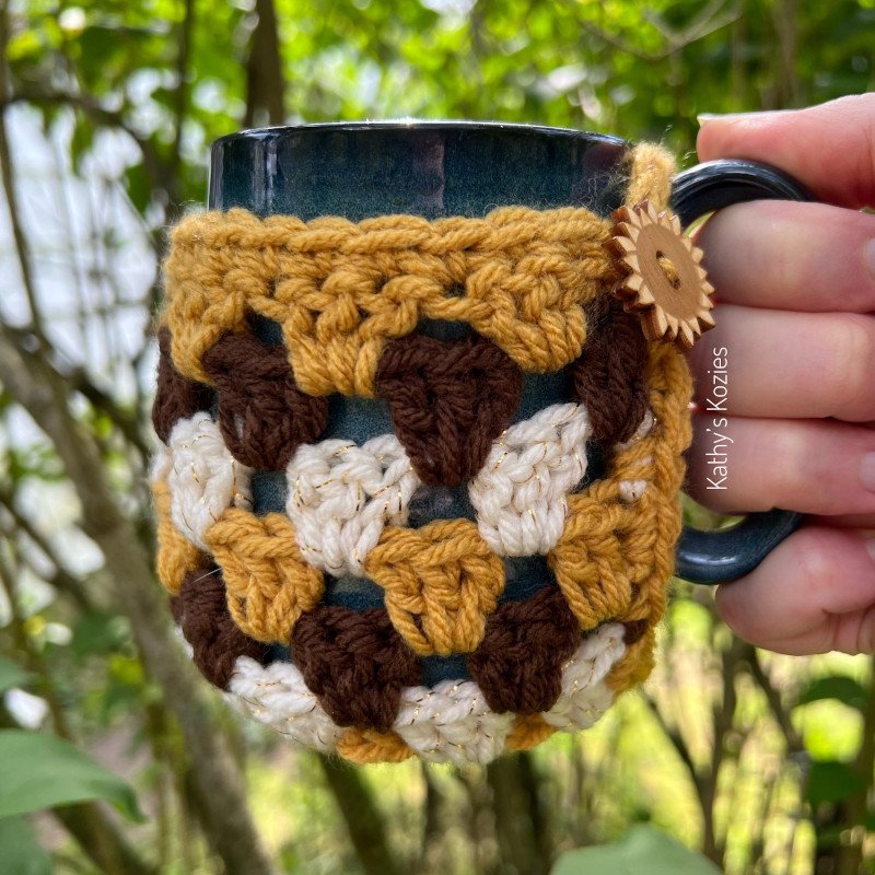 A right hand is holding up a dark blue/grey mug. There are trees and green leaves in the background. The mug is wrapped in a granny square cosy which is made with mustard, brown and cream yarn.