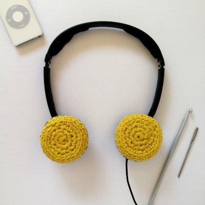 A pair of black headphones with the ear pieces covered in yellow crochet covers. The background is off white.