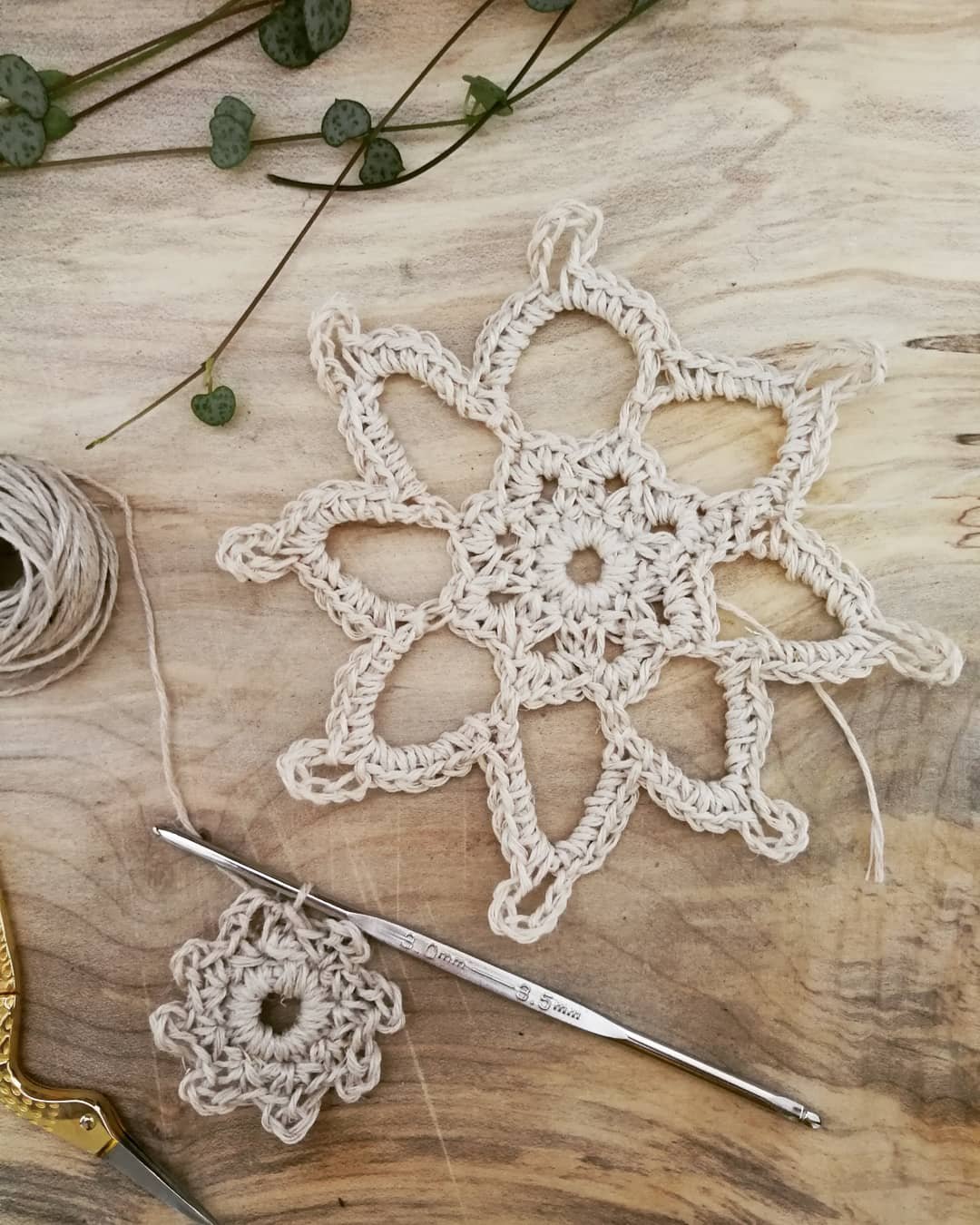 A natural coloured crocheted snowflake with 8 points is sat on a light wooden board. Each point has a triangular open space below it which leads onto a central flower shape with 8 petals. There is a small hole in the centre.
