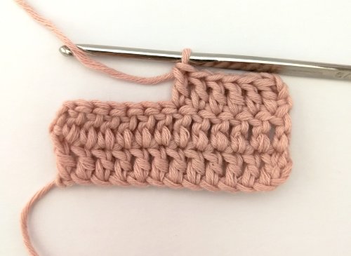 A small sample of crochet work worked in a pale pink yarn. There are two full rows of 12 double crochet stitches and half a third row. The working yarn is under the silver crochet hook and in the hook ready for the next stitch.