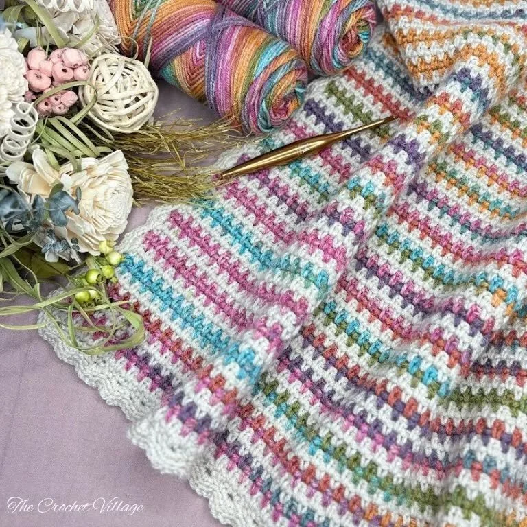A multicoloured throw is lying across a pale grey surface. The colours of the throw are mostly pastel colours with a cream base and border. The skeins of yarn and golden hook are laid next to the throw. Some white and pink flowers are in the top left