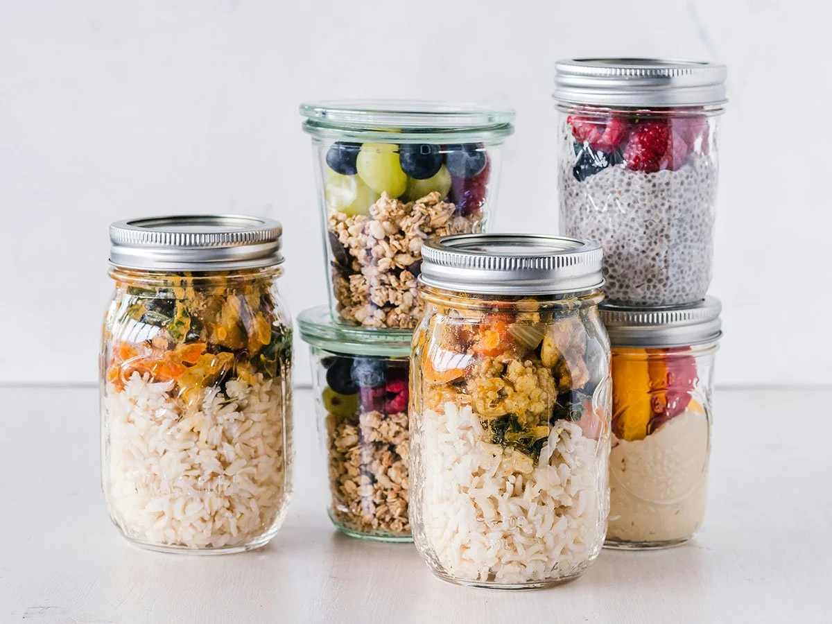 Six glass jars, four with silver lids, sat on an off white surface in front of a light grey background. The jars are filled with homemade food like rice and vegetables, chia pudding and granola with fruit.