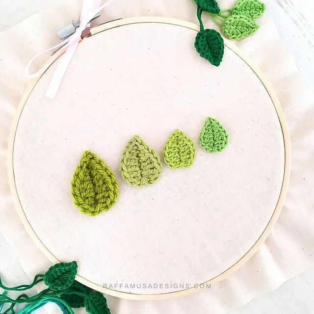 A birds eye view of a cross stitch hoop with a cream fabric. Four small leaves are centrally places from big to small, left to right in a slight diagonal. They are all different shades of green.